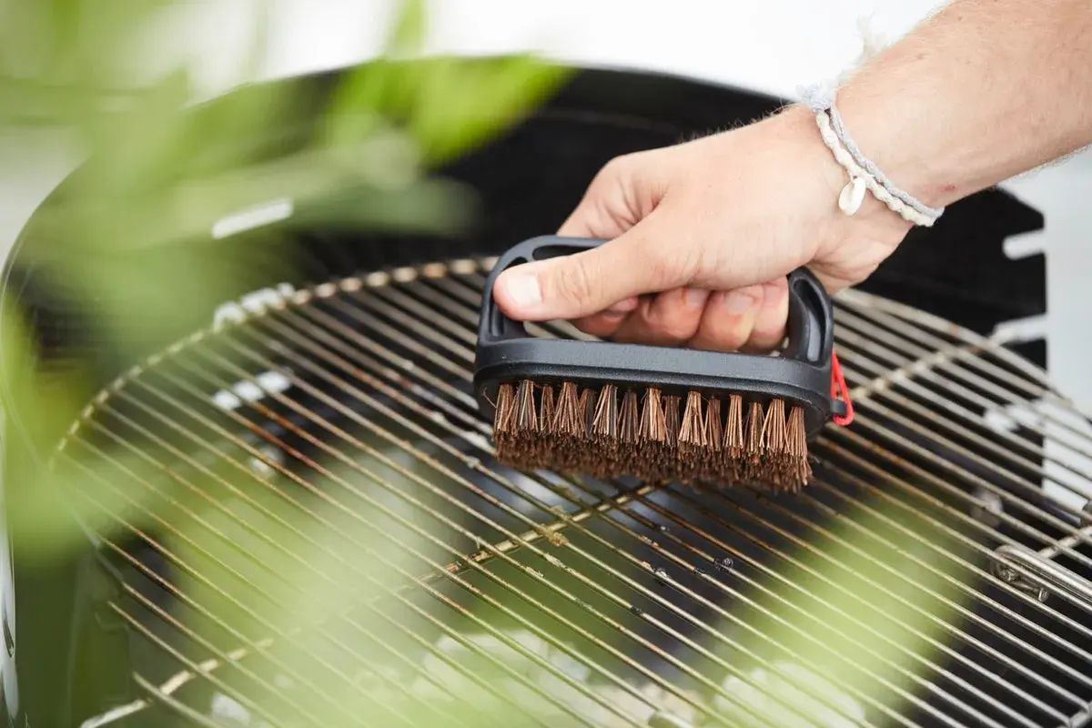 Main tenant une brosse pour nettoyer la grille du barbecue. Les herbes vertes floutées ajoutent une touche de fraîcheur à la scène.