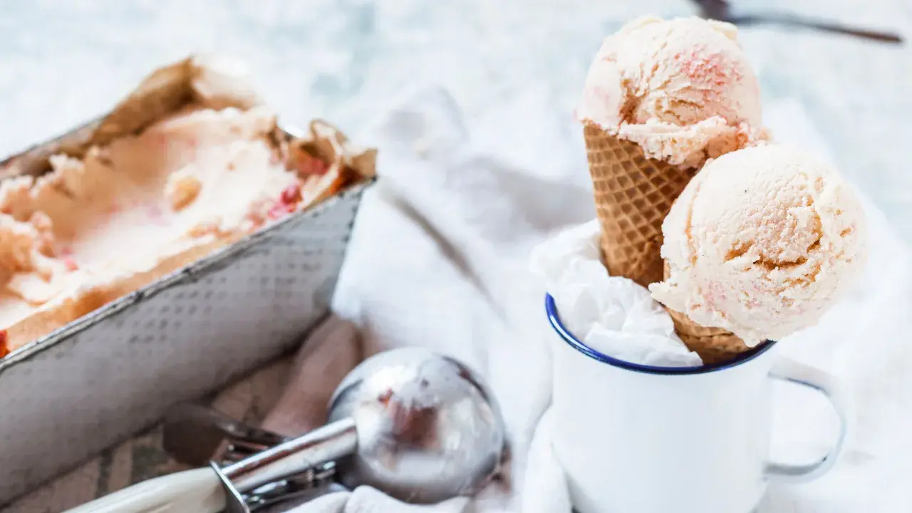 Due palline di gelato fatto in casa, con pezzetti di fragola, in un cono, accanto a una vaschetta e un porzionatore.