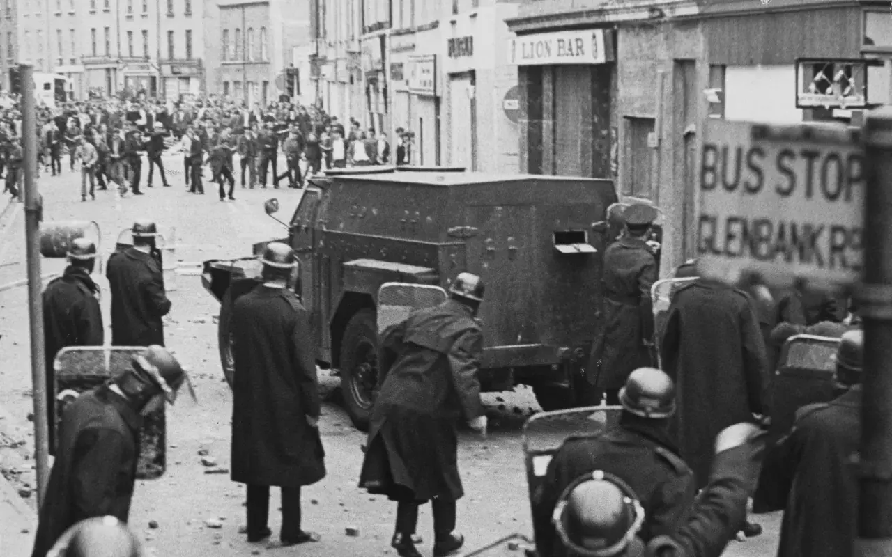 Unruhen in Nordirland, ein gepanzertes Fahrzeug und Polizisten mit Schilden. Die Szene erinnert an die Zeit vor dem Karfreitagsabkommen.