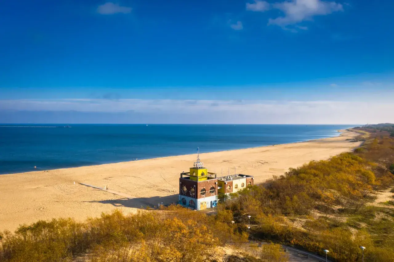 Piękna plaża i morze, idealne na spacer po jednej z **miejscowości nad morzem koło Gdańska**. W oddali widać charakterystyczny budynek.