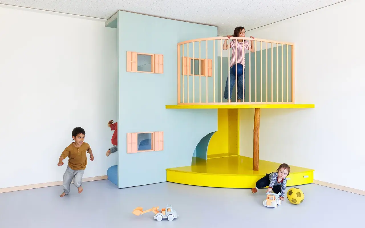 Kinder erkunden eine bunte Lernwerkstatt Kita. Ein Junge rennt, ein anderer versteckt sich, während ein Mädchen oben auf dem Spielturm steht.