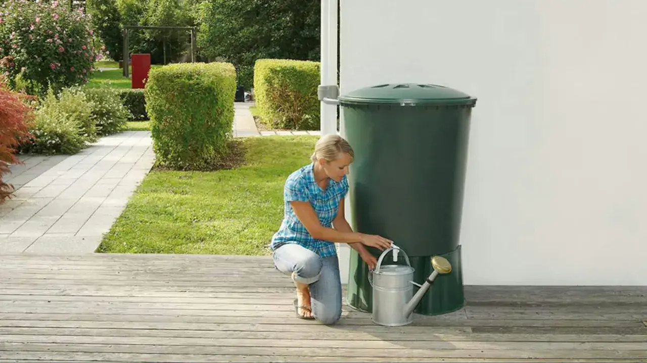 Donna raccoglie acqua piovana con un annaffiatoio da un serbatoio verde in giardino. Un modo ecologico per innaffiare le piante.