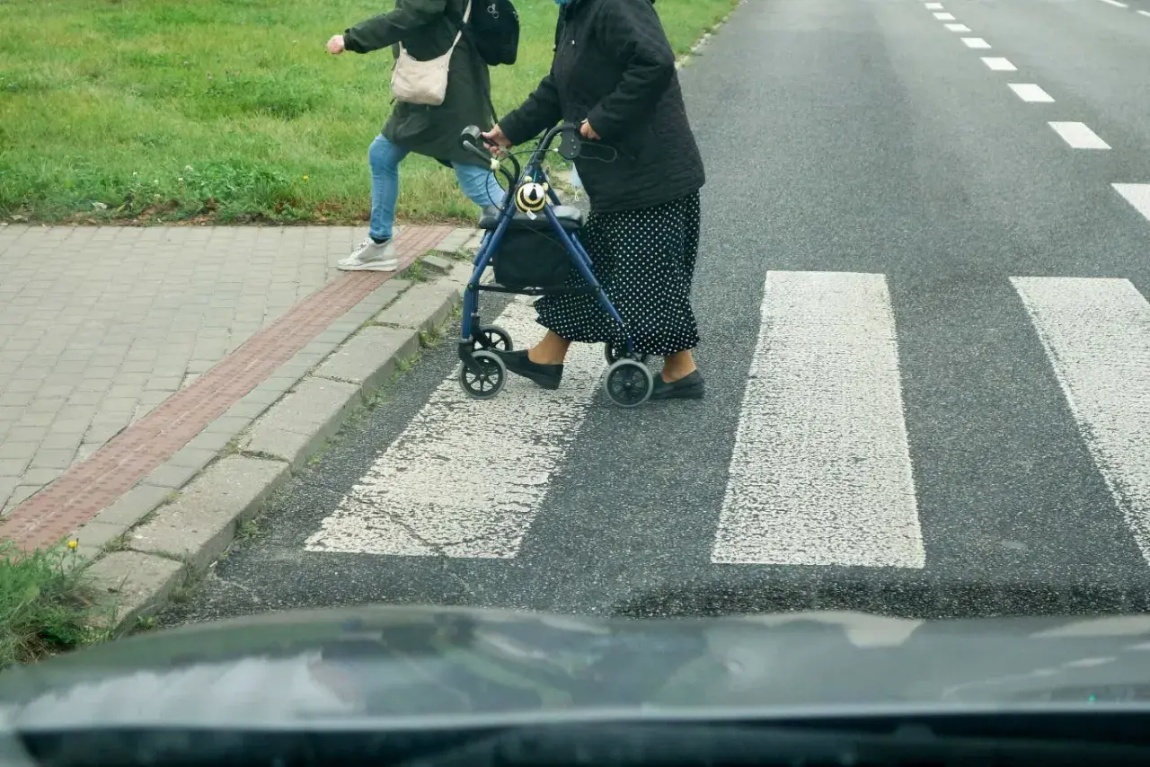 Starsza pani z chodzikiem i młodsza osoba przechodzą przez przejście dla pieszych.