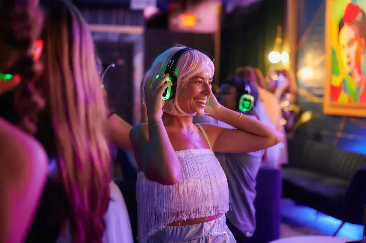 Uśmiechnięta kobieta z blond włosami cieszy się imprezą w stylu silent disco, zakładając świecące na zielono słuchawki.
