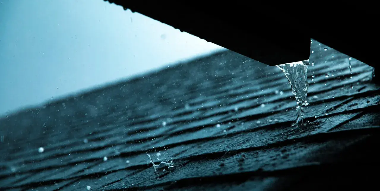 Regen prasselt auf ein dunkles Dach. Wasser tropft aus einer Rinne, ein Zeichen, dass das Blechdach reparieren nötig ist.