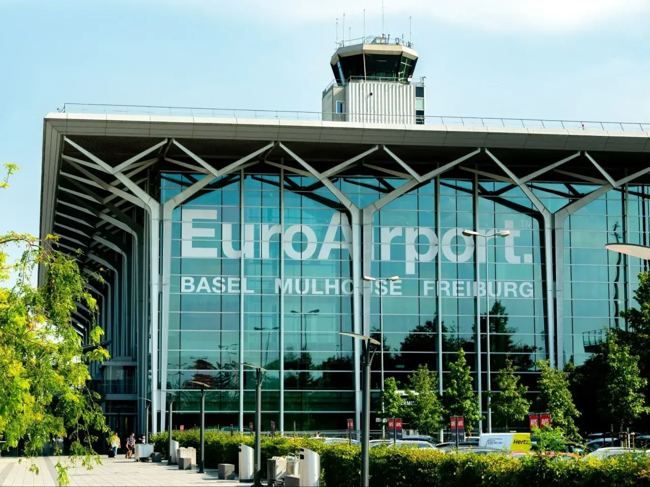 Nowoczesny budynek lotniska EuroAirport Basel Mulhouse Freiburg z charakterystyczną wieżą kontroli lotów i zielenią przed fasadą.