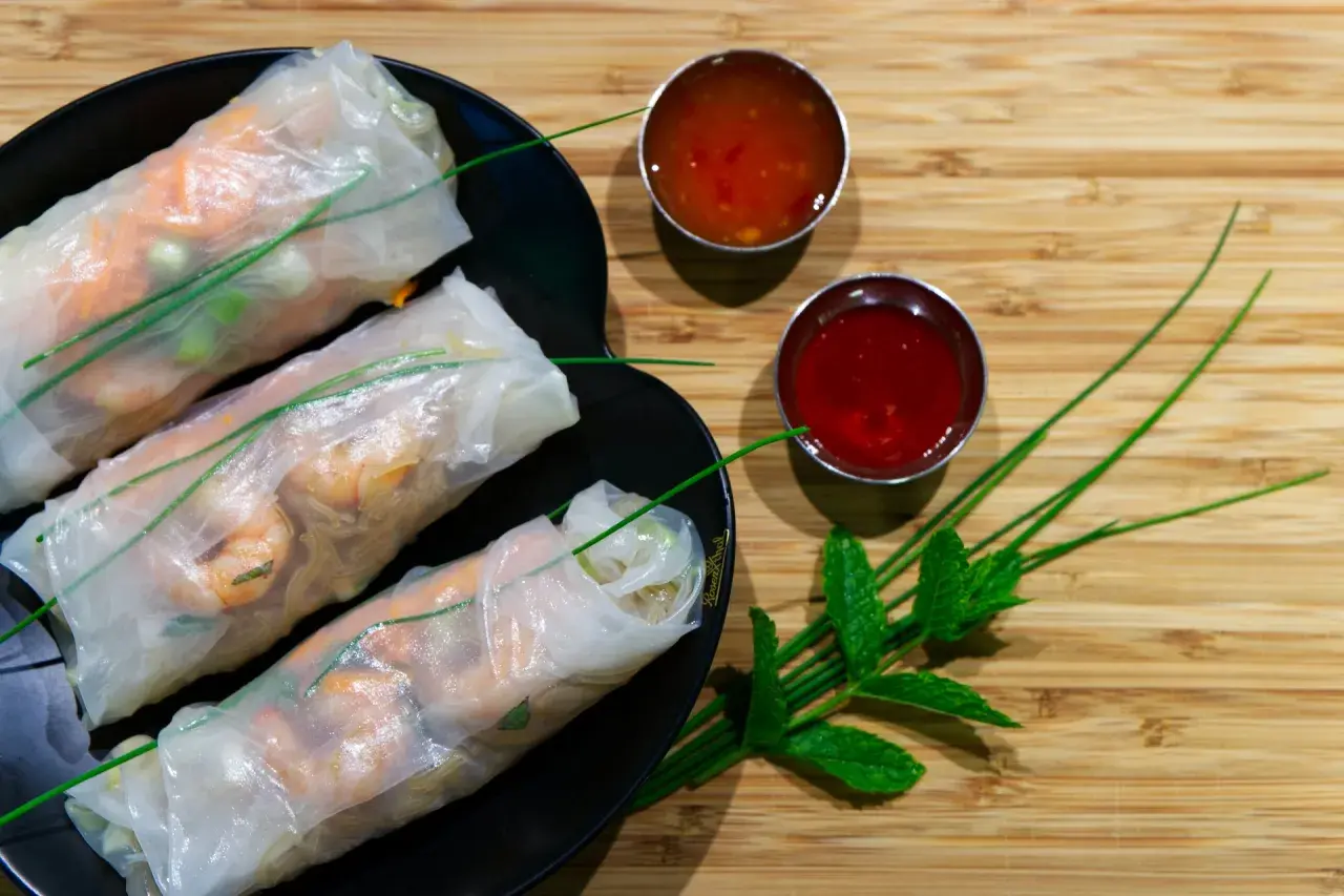 Tres rollitos de verano frescos con camarones y verduras, servidos en un plato negro con dos salsas y hojas de menta.