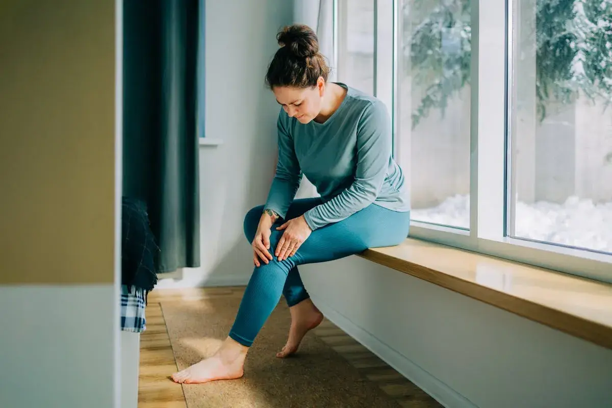 Femme assise près d'une fenêtre, tenant son genou, possiblement à cause d'une **durée courbature** après un effort.