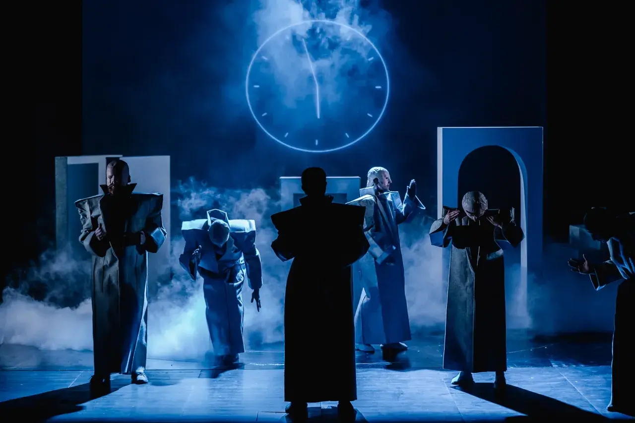 Postacie w futurystycznych strojach na scenie Teatru Arlekin w Łodzi. W tle świecący zegar.