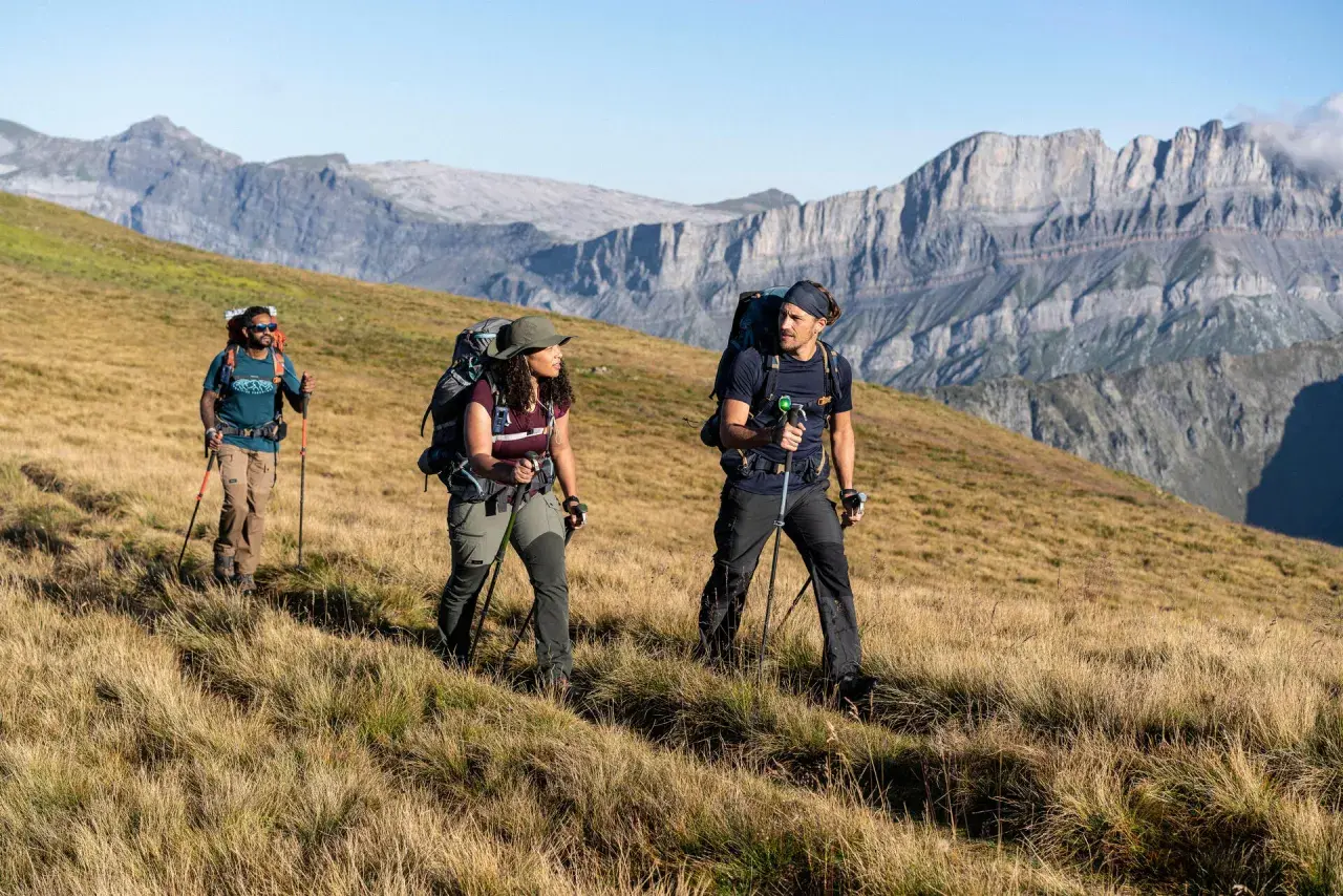 Trzech turystów z plecakami i kijkami przemierza górski szlak, przypominający **Pacific Crest Trail**.