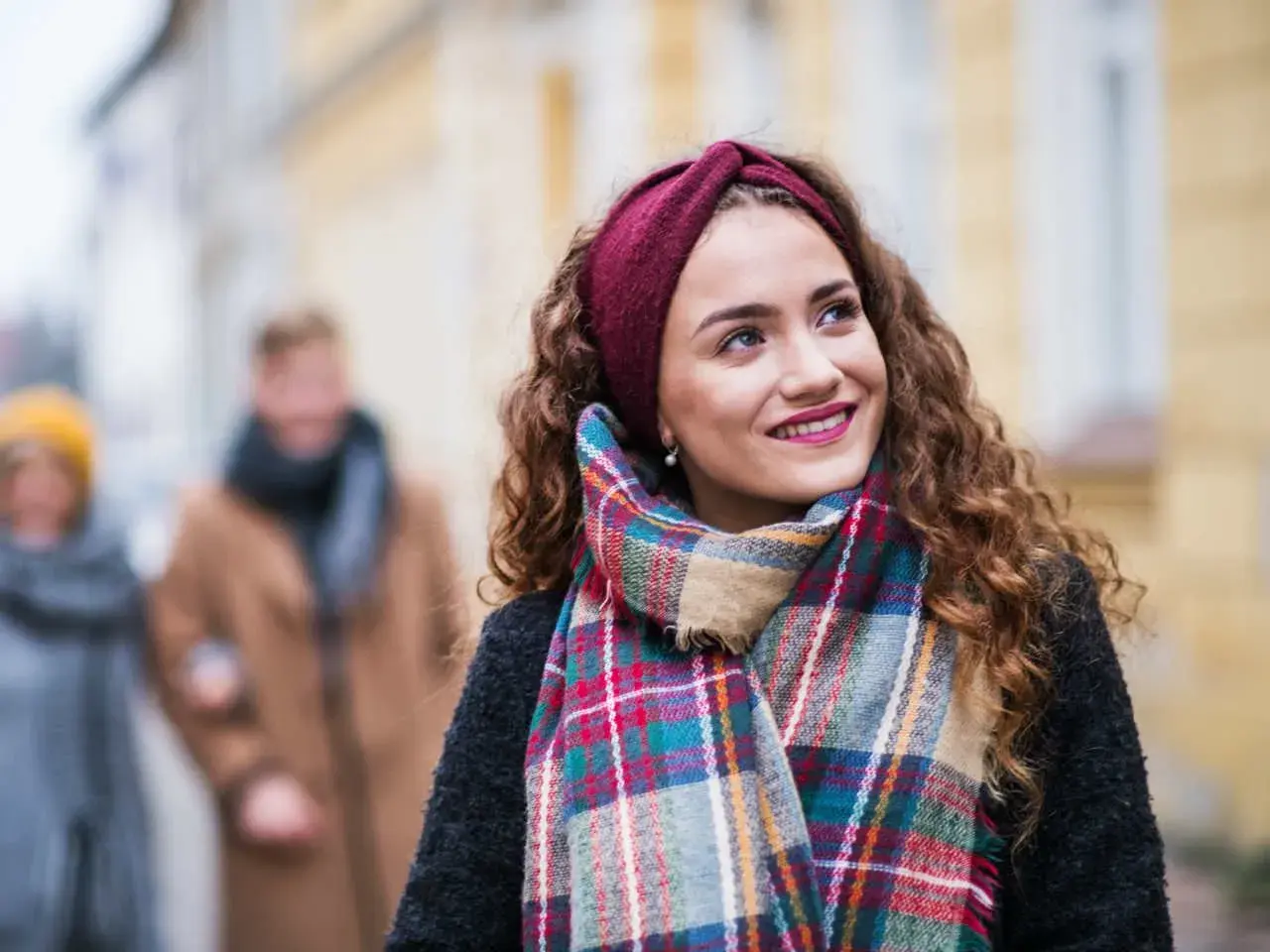 Uśmiechnięta dziewczyna w bordowej opasce i kolorowym szalu, idealnym do płaszcza w kratę. W tle para spacerująca ulicą.