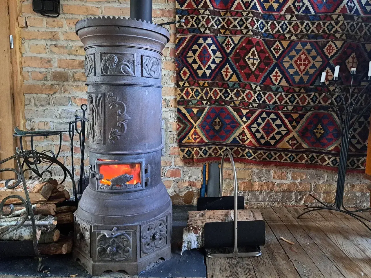 Stary, ozdobny kominek czy koza z płonącym ogniem, obok stos drewna i kolorowy kilim na ceglanej ścianie.