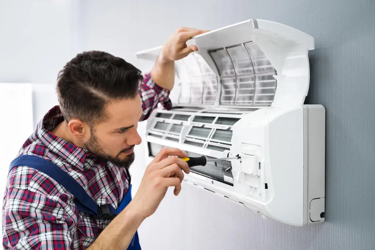 Technik z wkrętarką naprawia otwartą jednostkę klimatyzacyjną zamontowaną na ścianie.