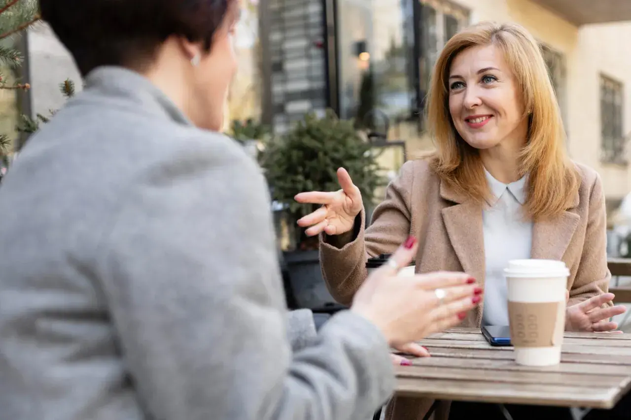 Dwie kobiety rozmawiają przy stoliku, gestykulując. Jedna z nich, z blond włosami, uśmiecha się, opowiadając o swoich przemyśleniach na temat granic w związku.