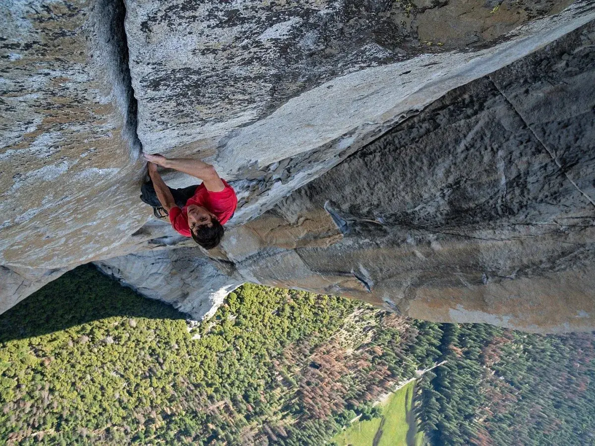 Alex Honnold klettert free solo die steile Felswand hinauf. Unter ihm erstreckt sich ein grünes Tal mit Bäumen.