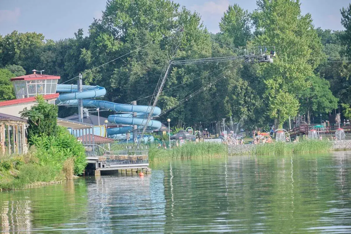 Letnia zabawa na **zalewie zemborzyckim**: niebieskie zjeżdżalnie wodne, ludzie kąpiący się i relaksujący się na plaży, otoczeni zielenią.