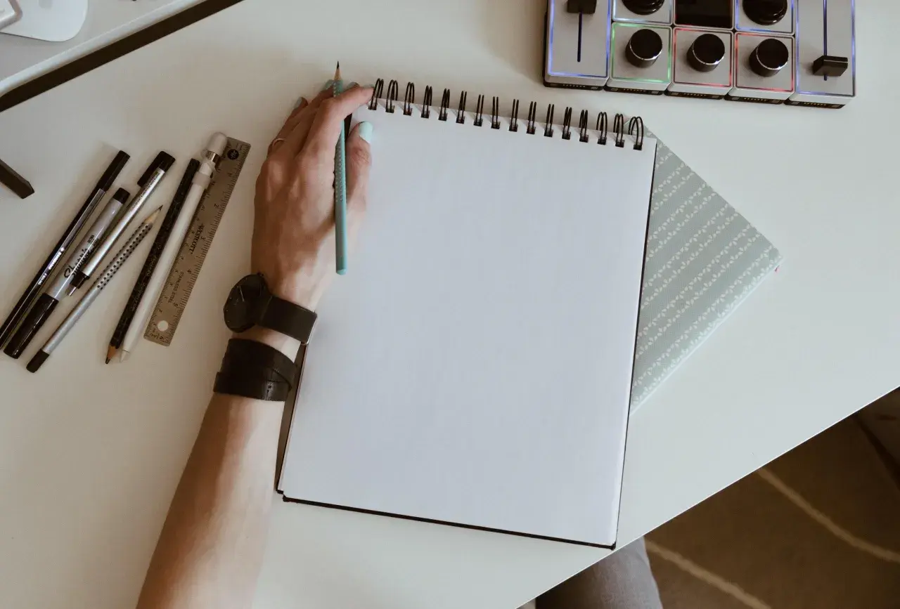 Mano con lápiz azul lista para crear dibujos de cumpleaños en un cuaderno blanco. A su lado, reglas, rotuladores y un reloj.