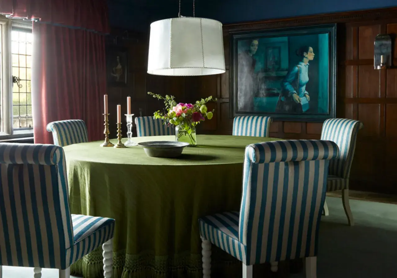 A dining room with a round green table, striped chairs, and a large painting. The table styles are classic and elegant.