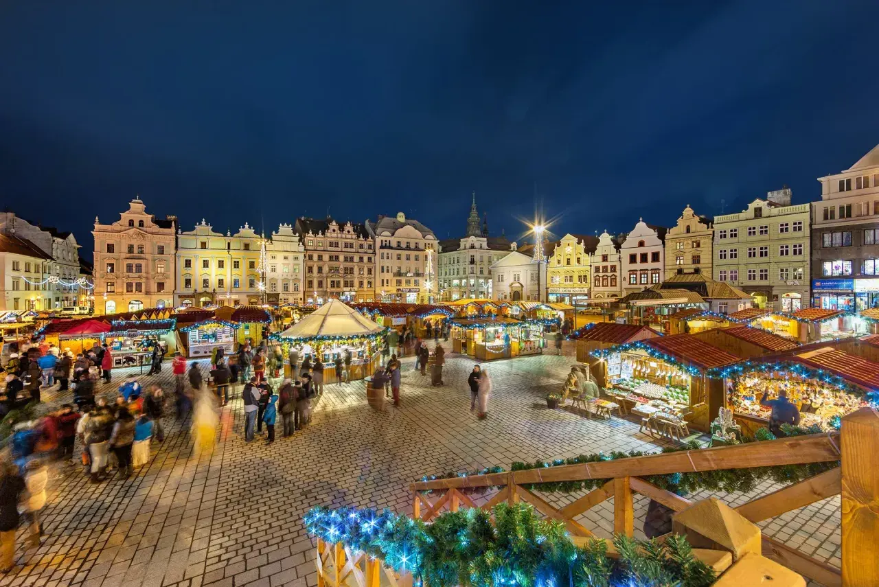 Beleuchtete Buden und historische Gebäude auf einem tschechischen Weihnachtsmarkt. Menschen schlendern über den Platz, der mit Lichterketten geschmückt ist.