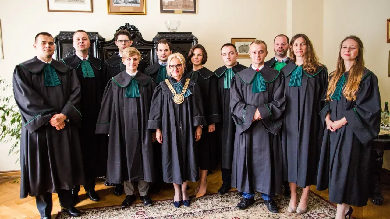 Grupa prawników w togach stoi w pomieszczeniu z meblami i obrazami.
