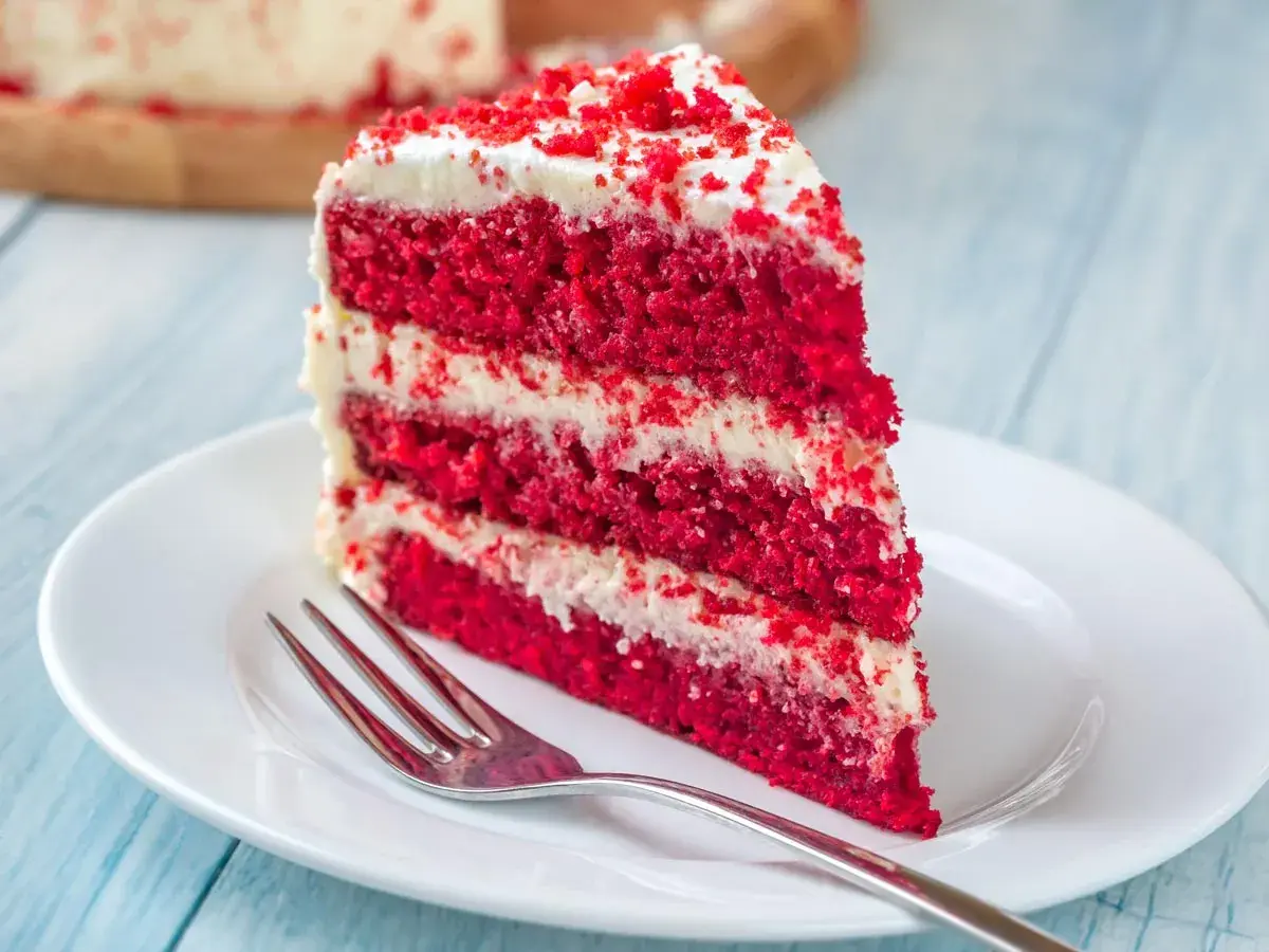 Un trozo de tarta red velvet con capas de bizcocho rojo y crema blanca, decorado con migas rojas. Un tenedor plateado espera en el plato blanco.
