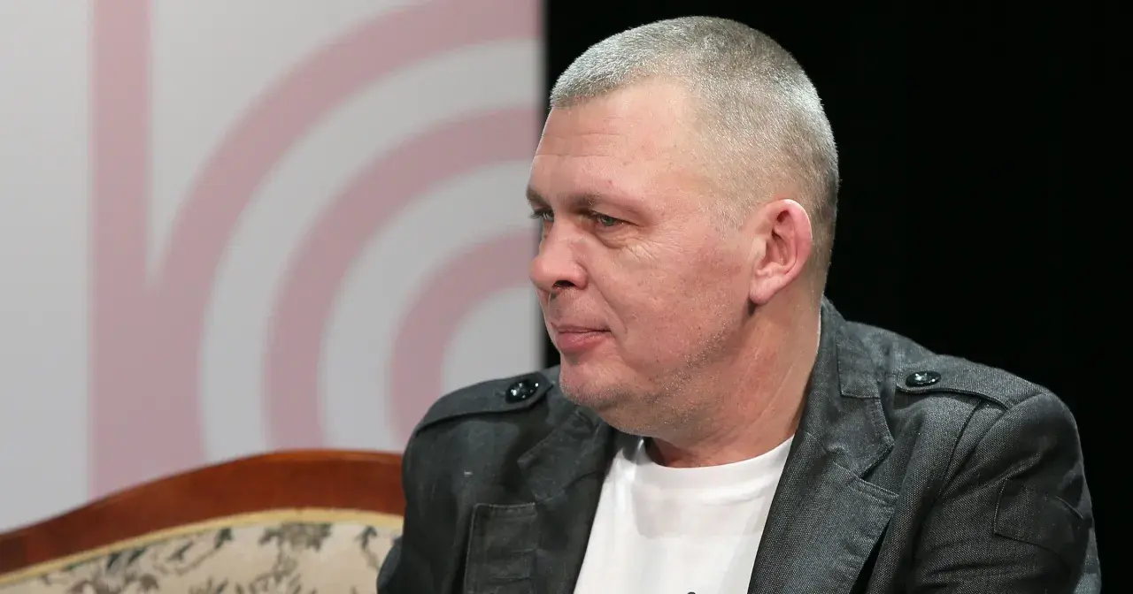 Mężczyzna z krótkimi włosami, w szarej kurtce, siedzi na kanapie. To wokalista zespołu Raz Dwa Trzy.