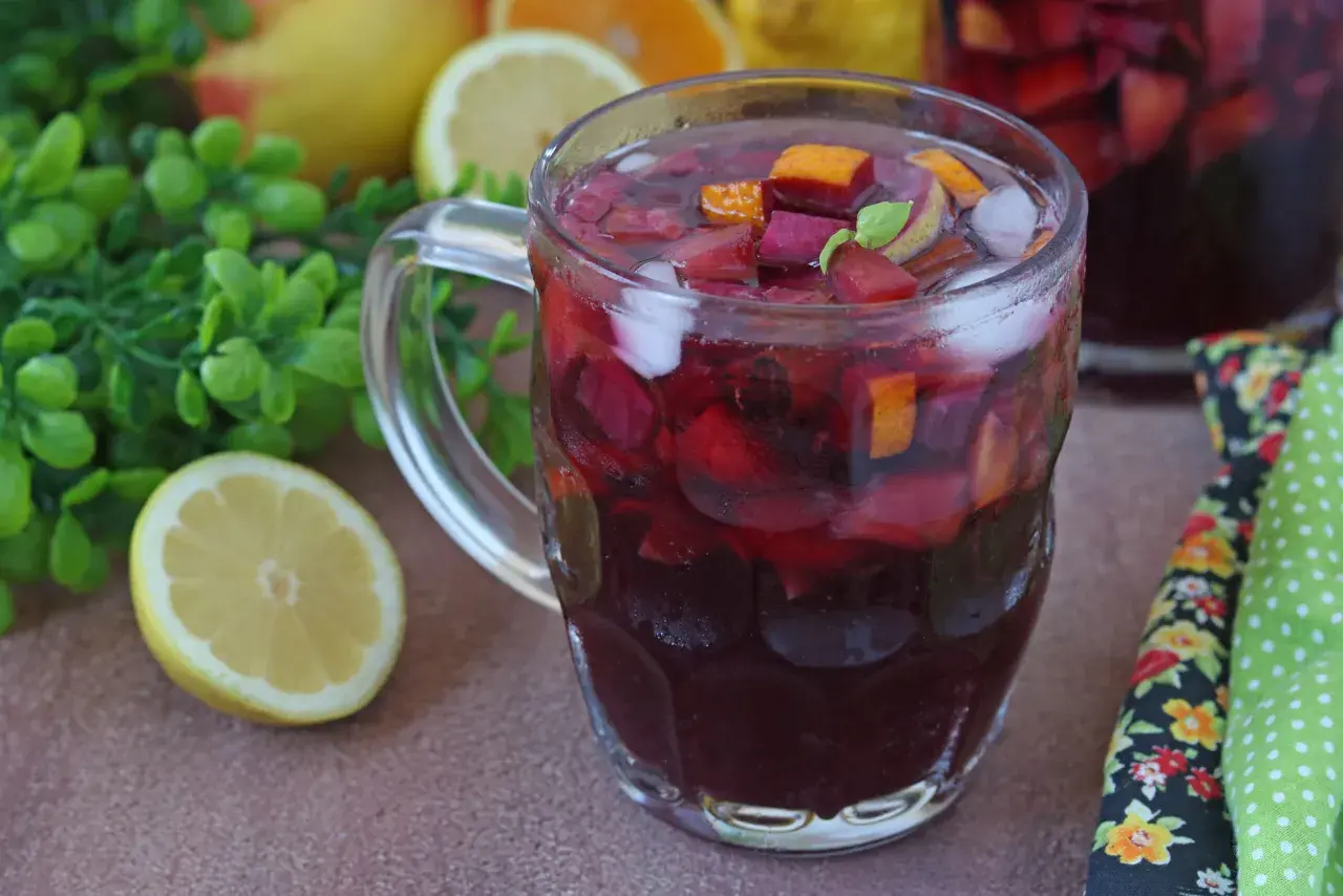 Un bicchiere colmo di sangria, con pezzi di frutta rossa, arancia e ghiaccio. La ricetta perfetta per l'estate, con limoni e foglie verdi sullo sfondo.