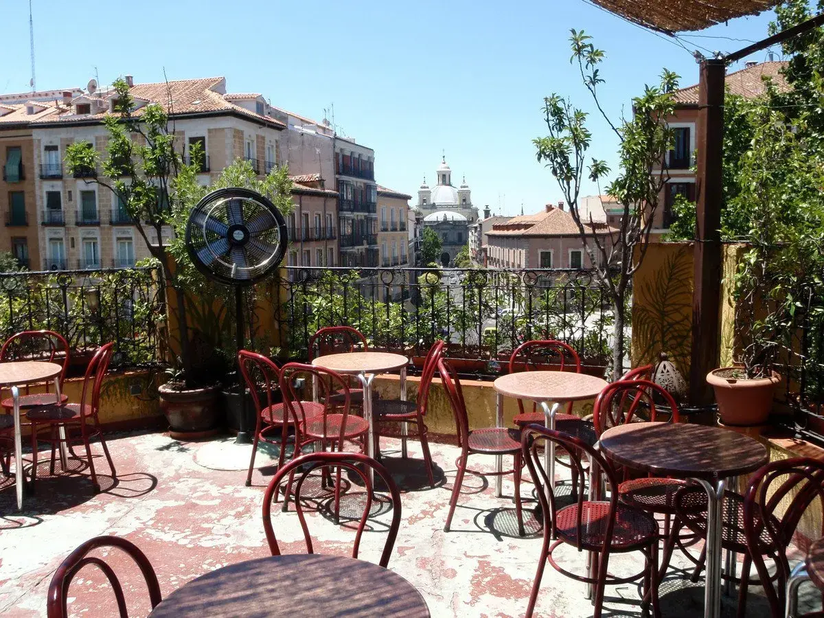 Terraza de verano en Madrid, con mesas y sillas rojas, ventilador y vistas a edificios históricos. Un lugar perfecto para el viajero madrileño.