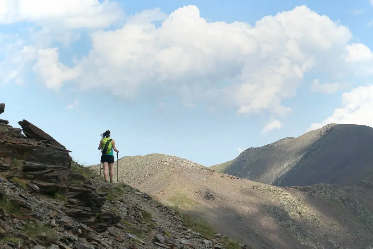 Senderista en la ruta Vall de Núria, con mochila y bastones, ascendiendo por un sendero rocoso con montañas al fondo.