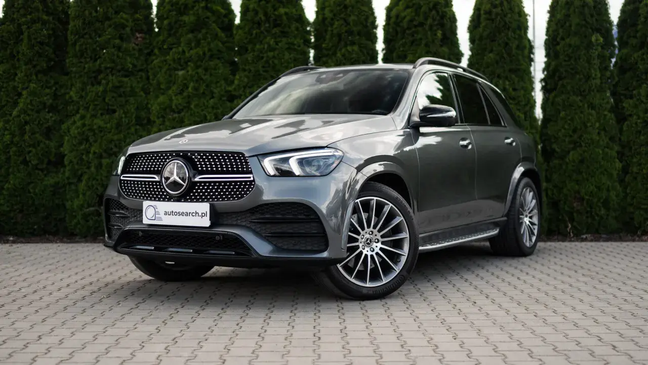 Elegancki, szary Mercedes GLE 400d SUV stoi na wybrukowanym podjeździe, otoczony gęstymi, zielonymi żywopłotami.