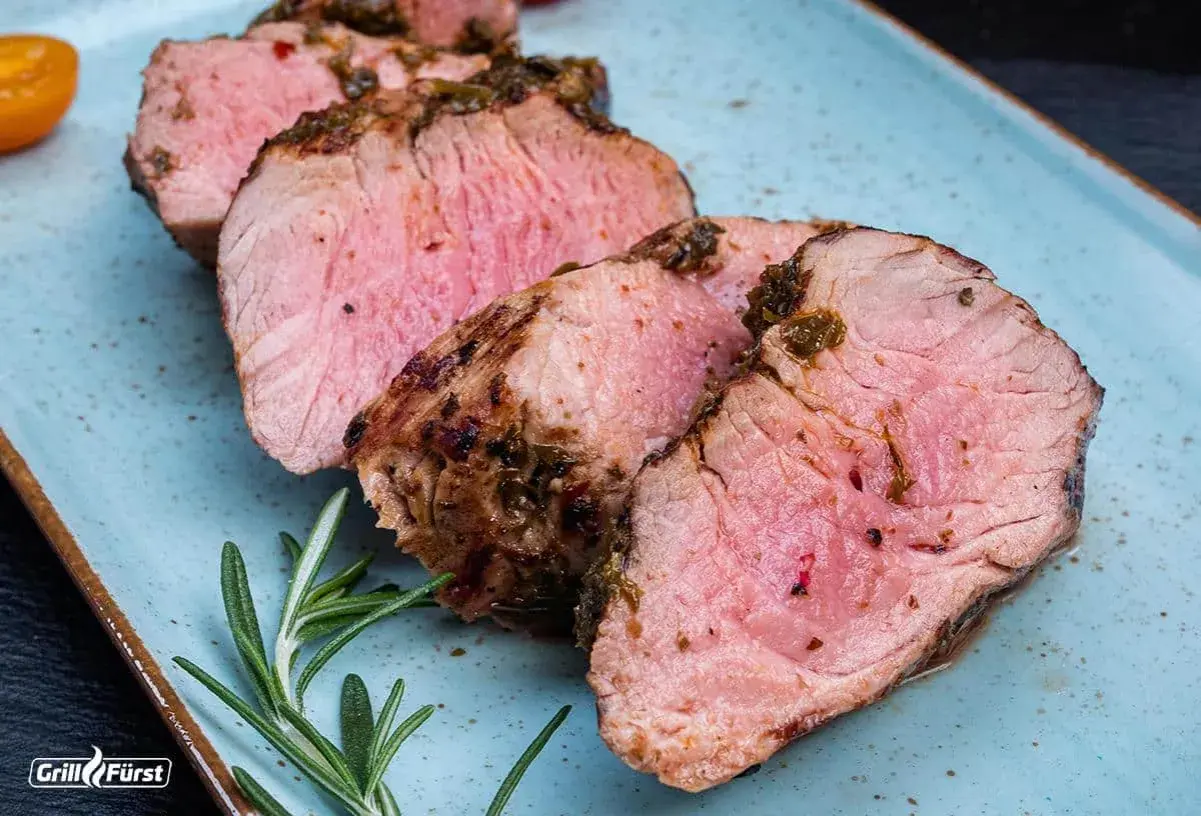 Saftiges schweinefilet grillen, perfekt rosa gebraten, mit Kräuterkruste und Rosmarin auf blauem Teller.
