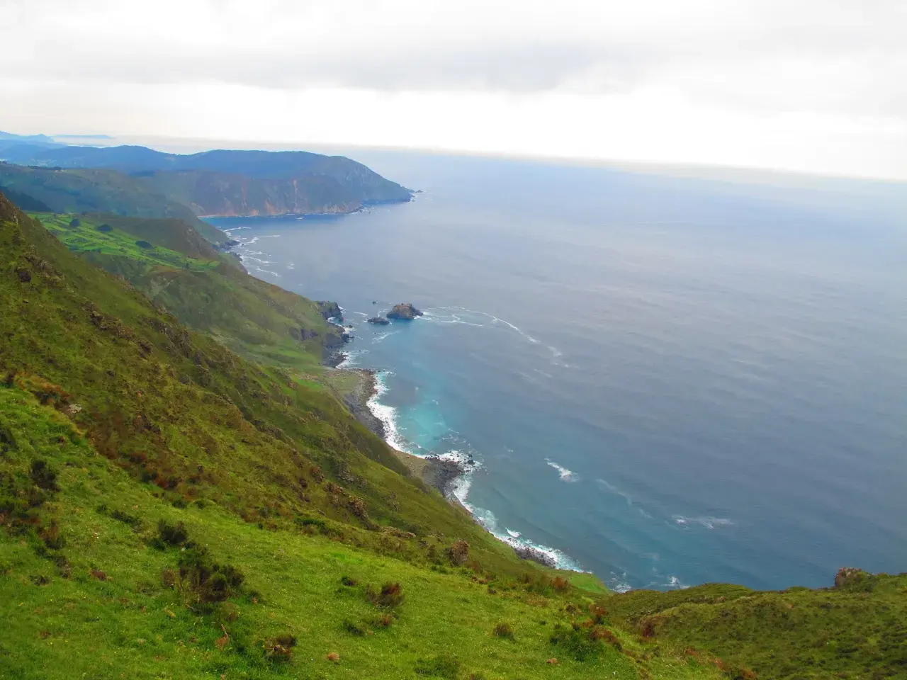 Paisaje costero verde y escarpado cerca de la garita de Herbeira, con el mar azul y las olas rompiendo en la costa rocosa.