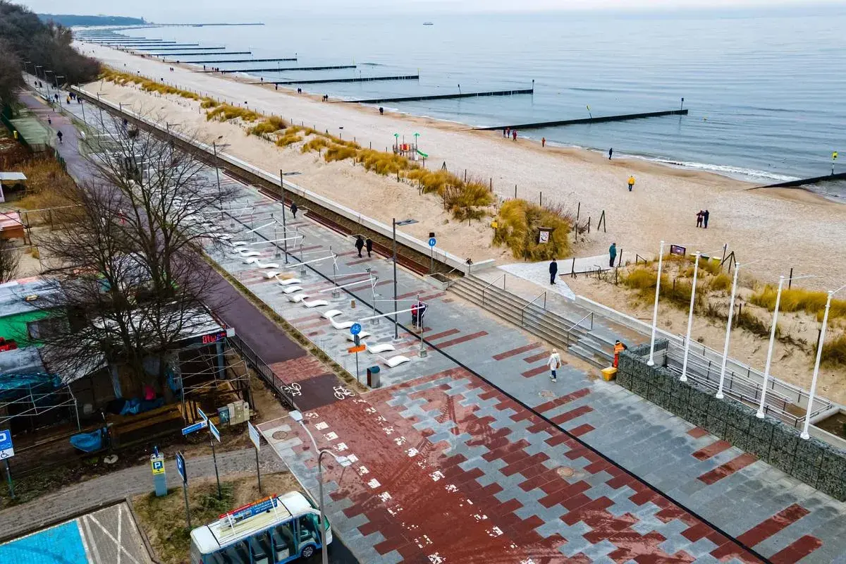 Nowoczesna promenada nad morzem, z piaskiem, wydmami i łagodnymi falami. Ludzie spacerują, ciesząc się chłodnym dniem.