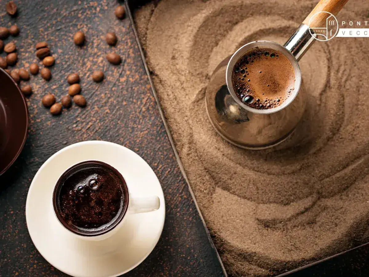 Un caffè arabo fumante in una tazzina bianca, accanto a un bricco di metallo su sabbia calda. Chicchi di caffè sparsi.