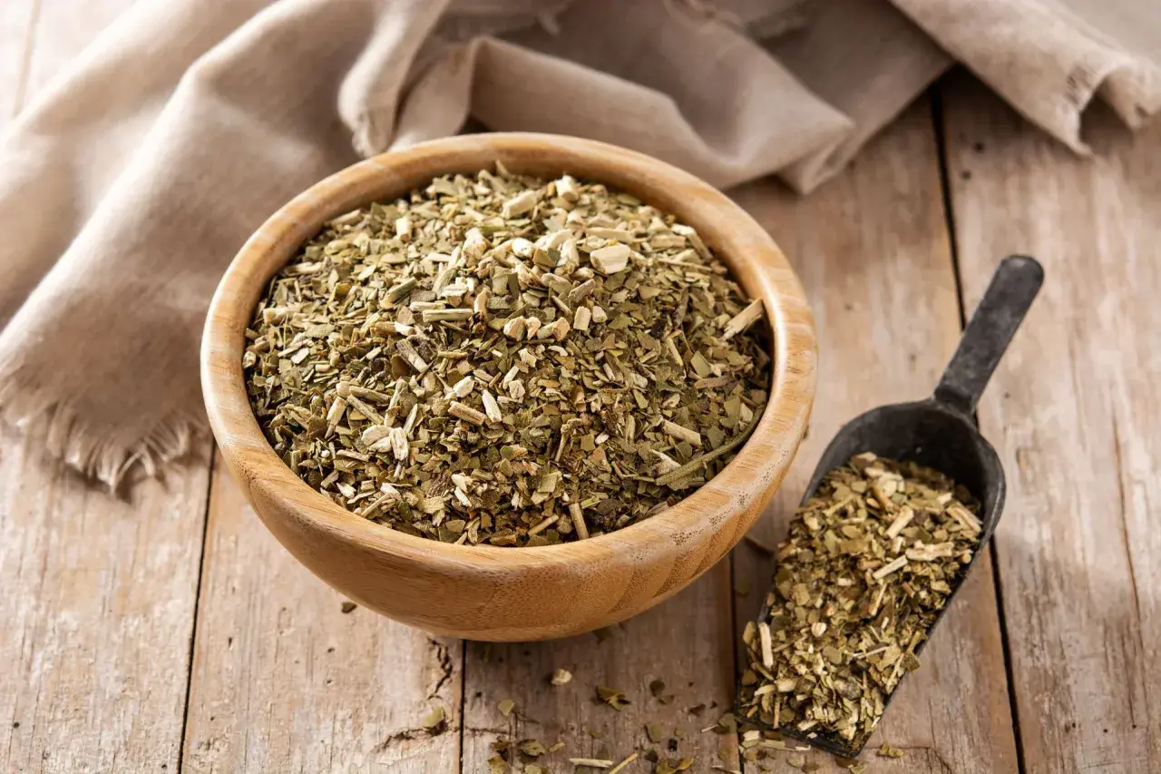 Suszone liście yerba mate w drewnianej misce i szufelce. Choć yerba mate ma wiele zalet, warto pamiętać o jej negatywnych właściwościach, np. o zawartości kofeiny.