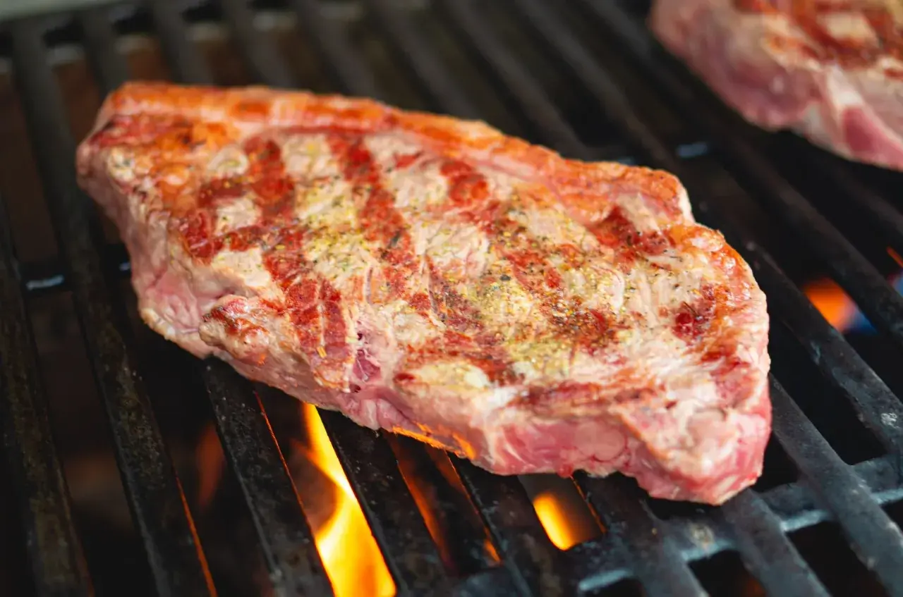 Un steak de basse côte, assaisonné, grésille sur un barbecue. Les flammes dansent sous la grille, promettant une cuisson parfaite.