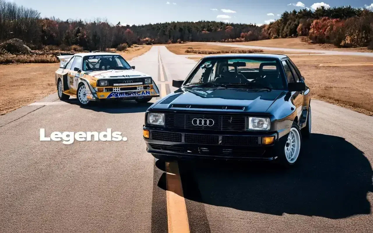 Dwa kultowe Audi S1 Quattro, jeden czarny, drugi w barwach rajdowych, stoją na pustej drodze.