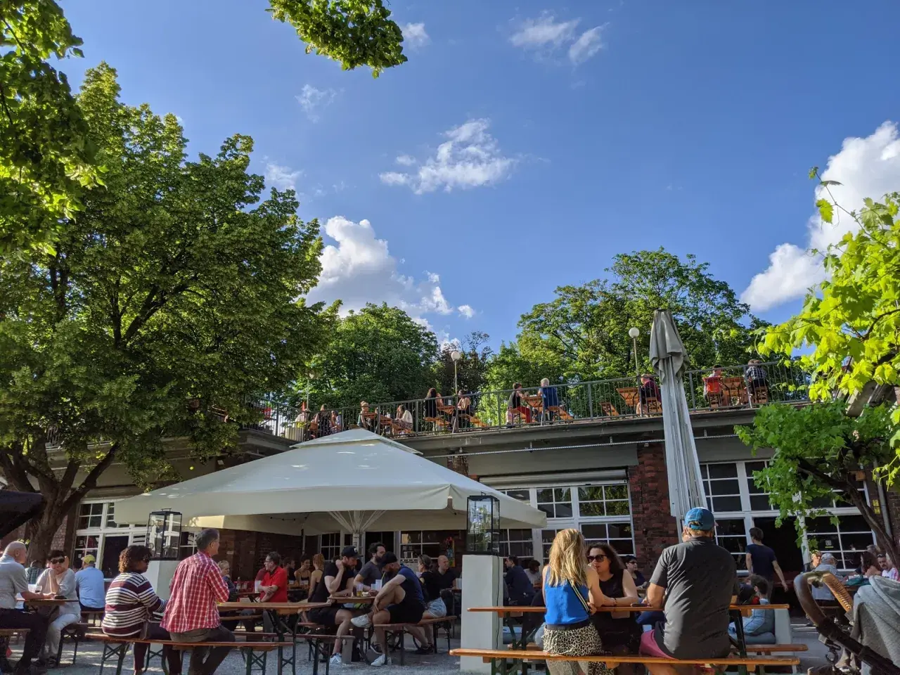 Menschen genießen das Wetter im Golgatha Biergarten. Sonnenschein, grüne Bäume und ein blauer Himmel bilden die Kulisse.