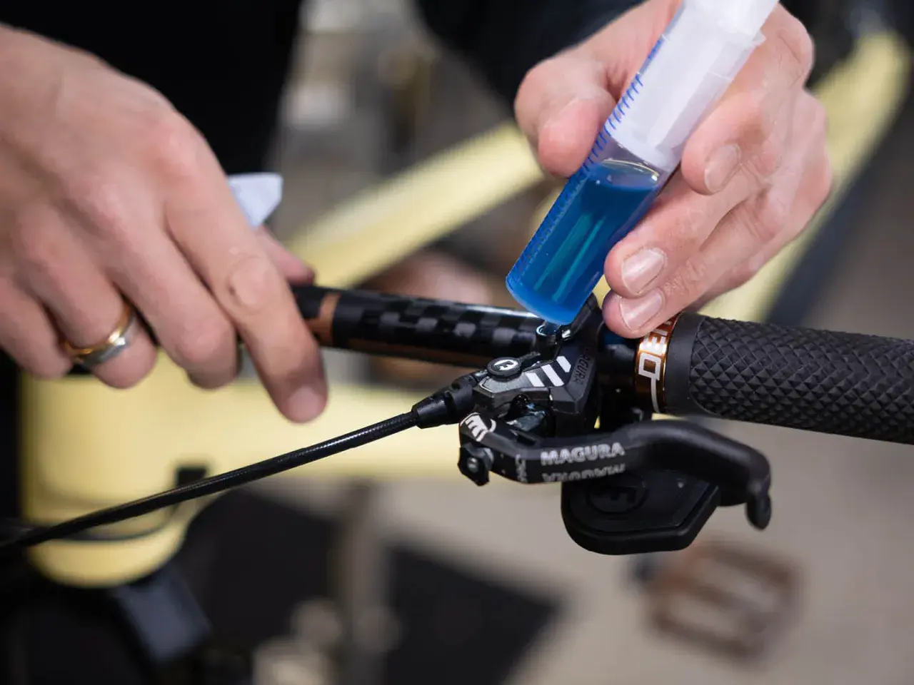 Hands add blue brake fluid from a syringe to a bicycle brake lever. This fluid is corrosive due to its hygroscopic nature, absorbing moisture.
