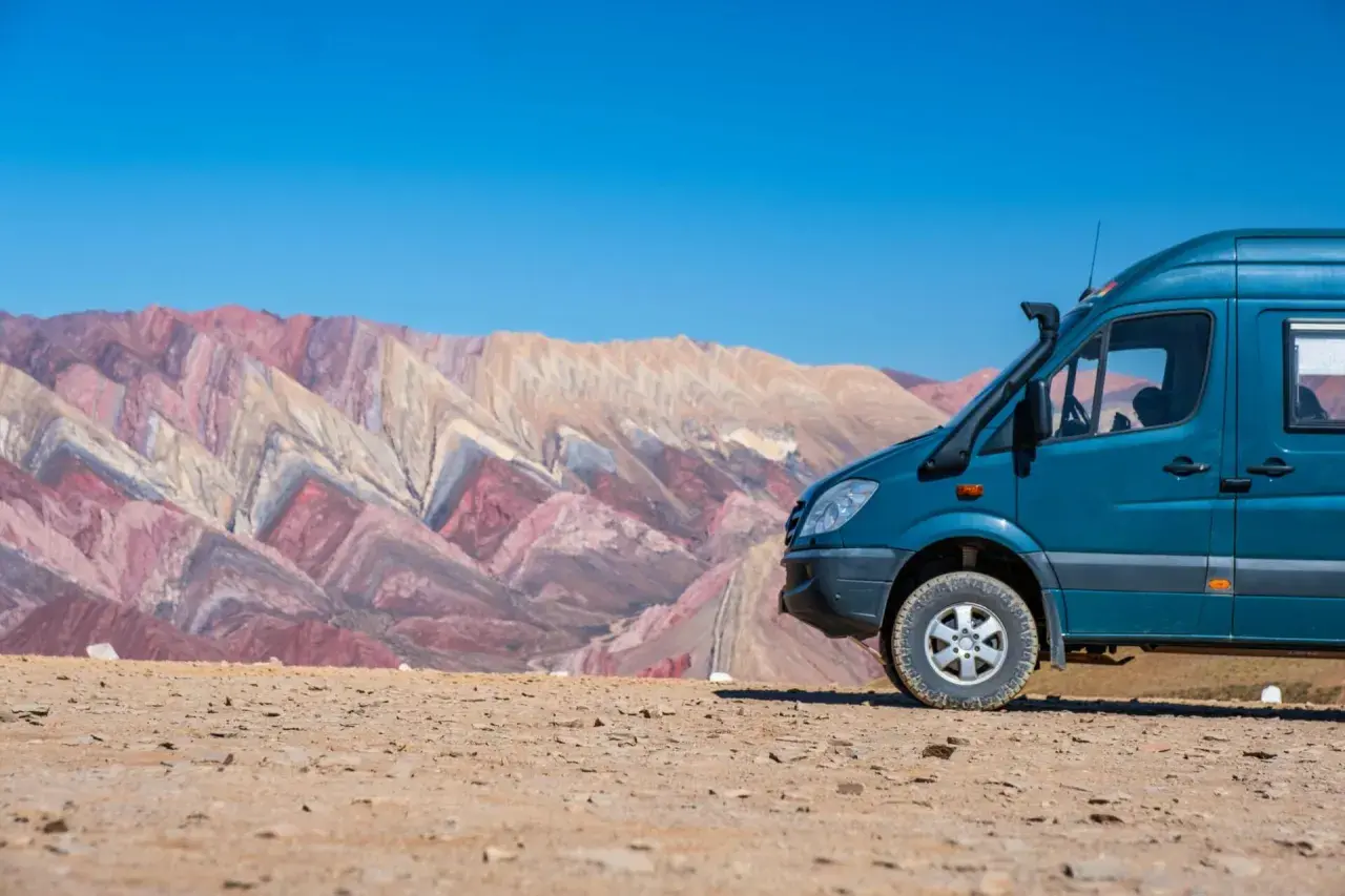 Un camping car tout terrain bleu est garé sur un sol rocailleux, avec en arrière-plan des montagnes aux couleurs vives et un ciel bleu éclatant.