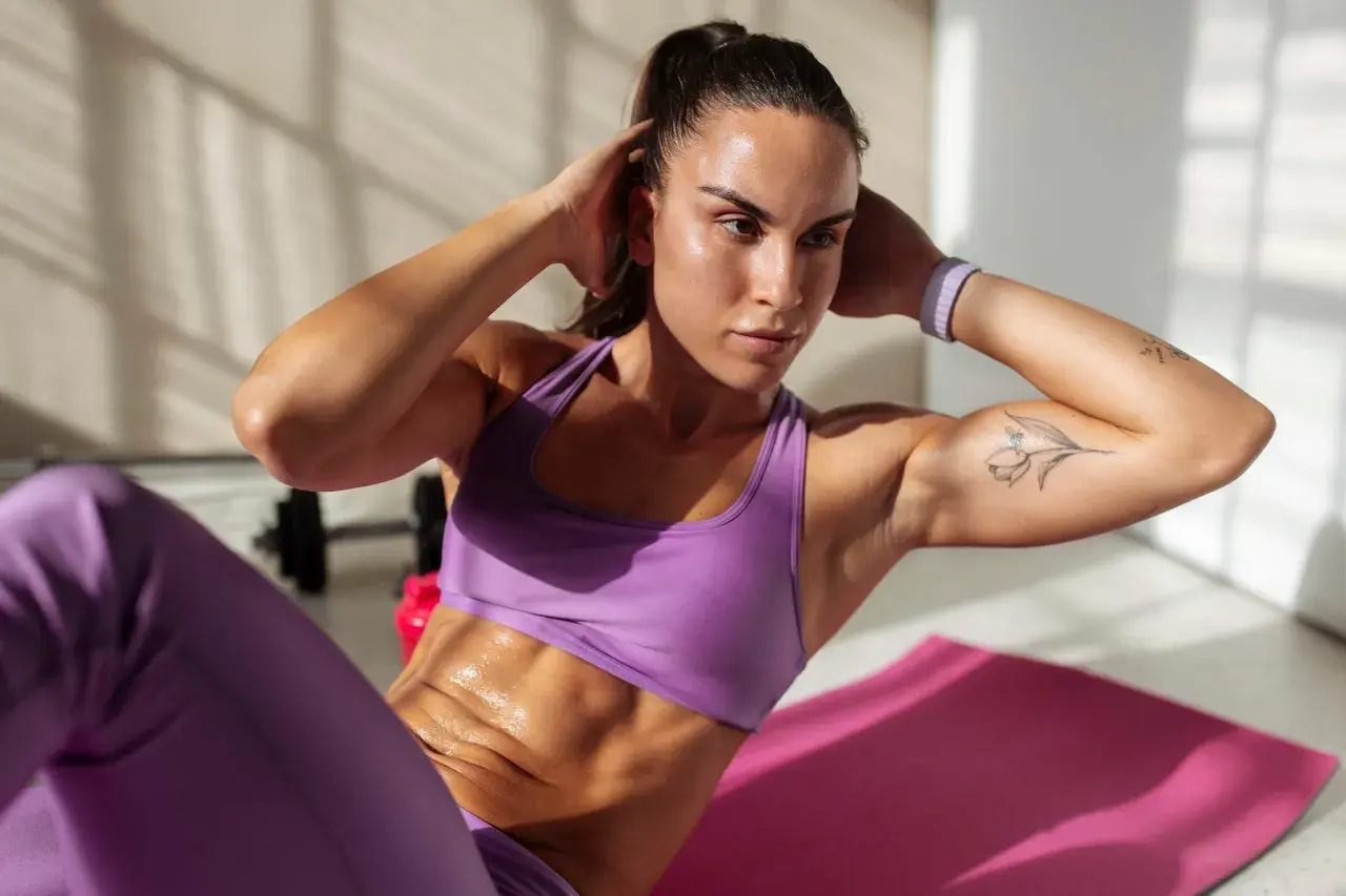 Frau macht Crunches auf einer rosa Matte, um einen flachen Bauch zu bekommen. Sie trägt lila Sportkleidung und hat ein Tattoo auf dem Arm.