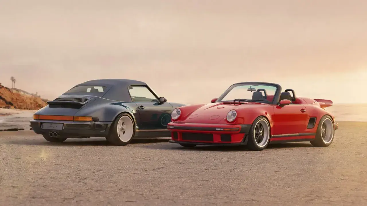Dos Porsche descapotables, uno rojo y otro azul oscuro, posan en la playa. El **Singer Porsche** rojo destaca con su diseño deportivo y el azul con su capota puesta.