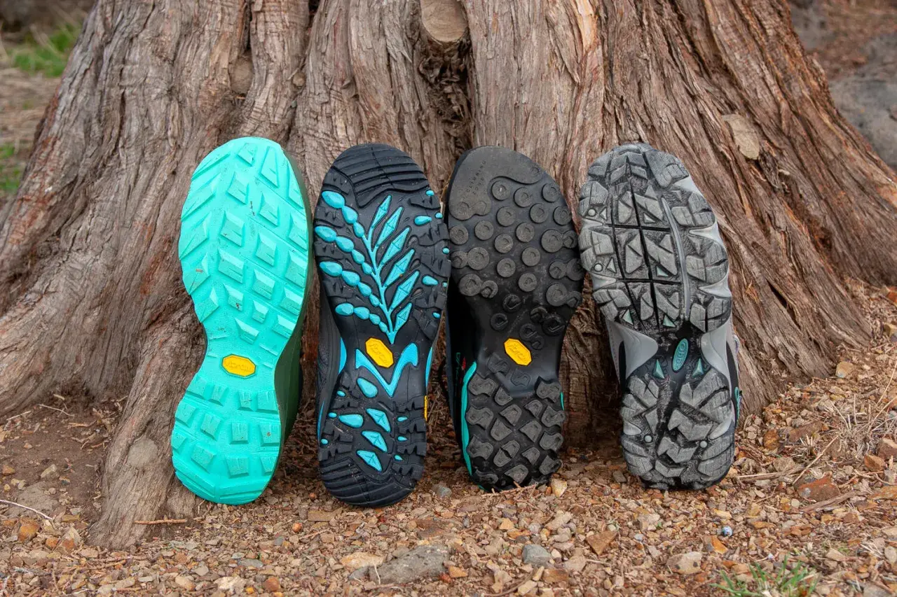 Four hiking boot soles lean against a tree. From left to right: bright teal, black with teal accents, black with circular lugs, and grey with a rugged tread. These sneaker soles are hard and slippery, designed for grip.