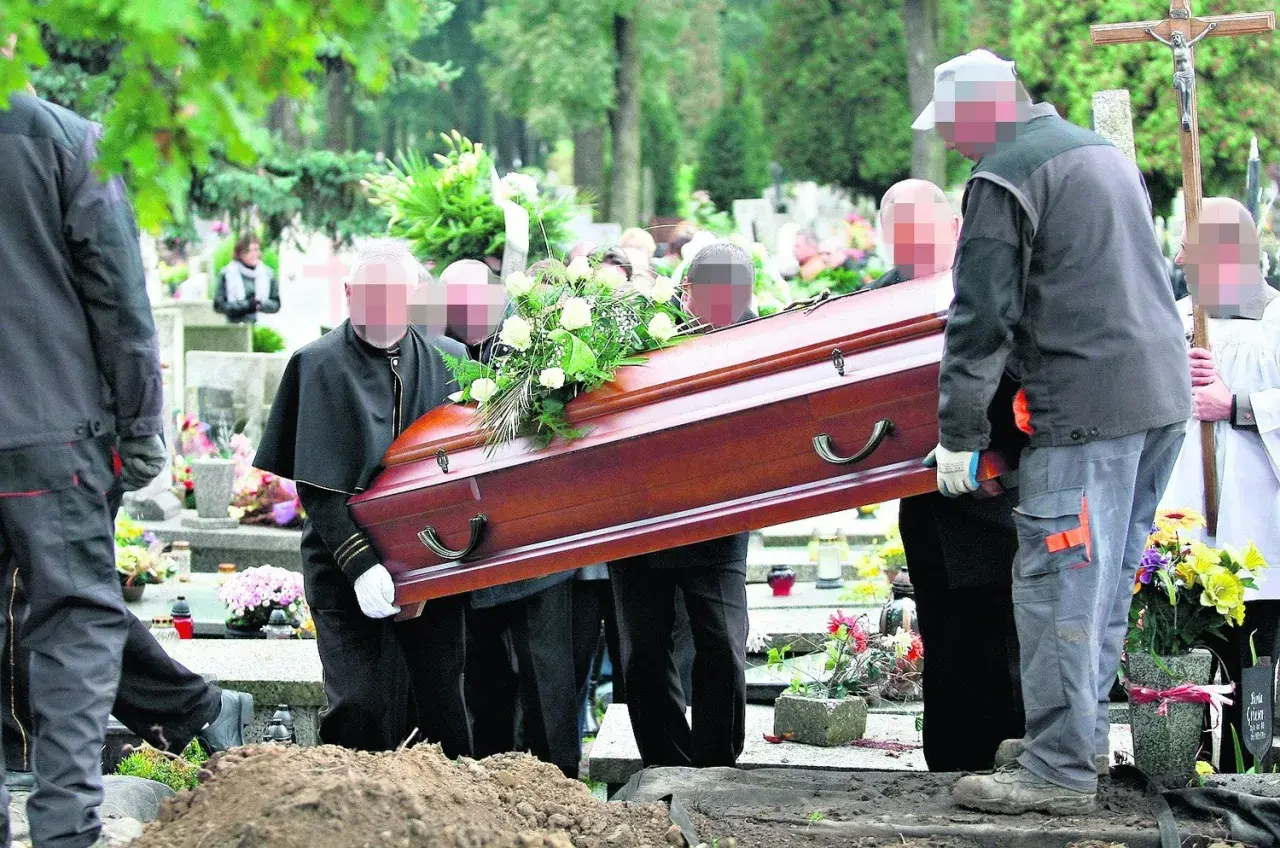 Pogrzeb na cmentarzu. Kilku mężczyzn niesie drewnianą trumnę ozdobioną kwiatami w kierunku grobu głębinowego.