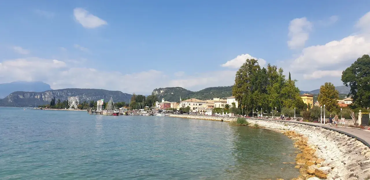 Il lungolago di Bardolino, con il lago azzurro, le barche, le case colorate e le montagne sullo sfondo. Una ruota panoramica si staglia all'orizzonte.