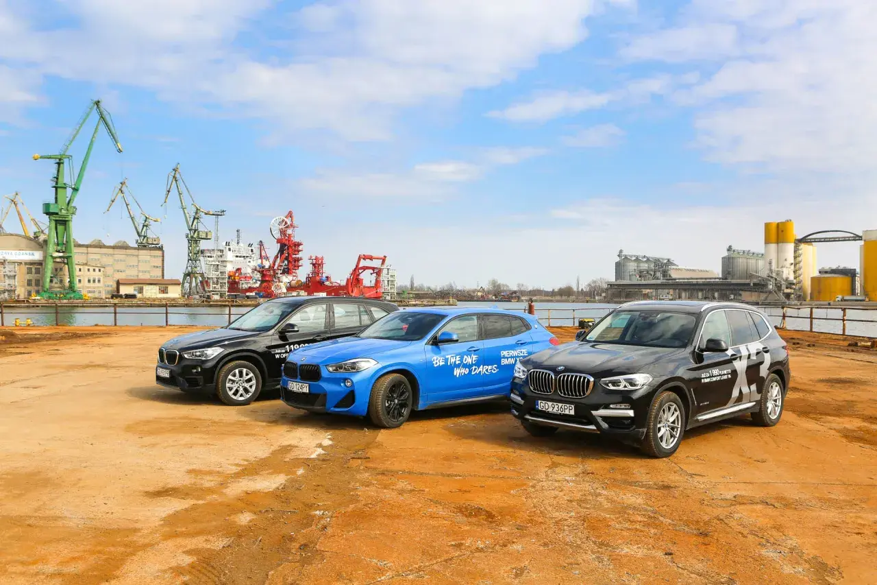 Trzy samochody BMW X: czarny, niebieski i kolejny czarny, stoją na placu budowy z dźwigami w tle.