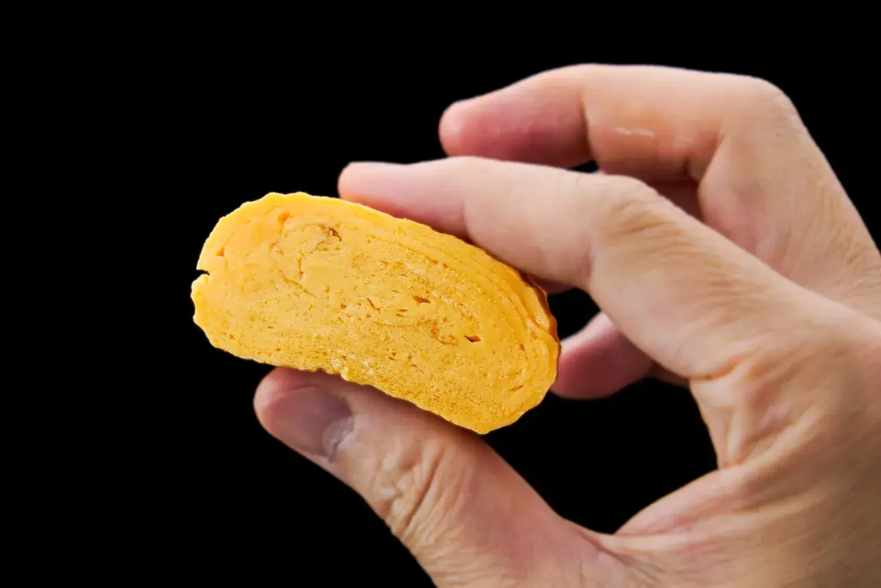 A hand holds a slice of golden-yellow dashimaki tamago, revealing its layered, fluffy texture.