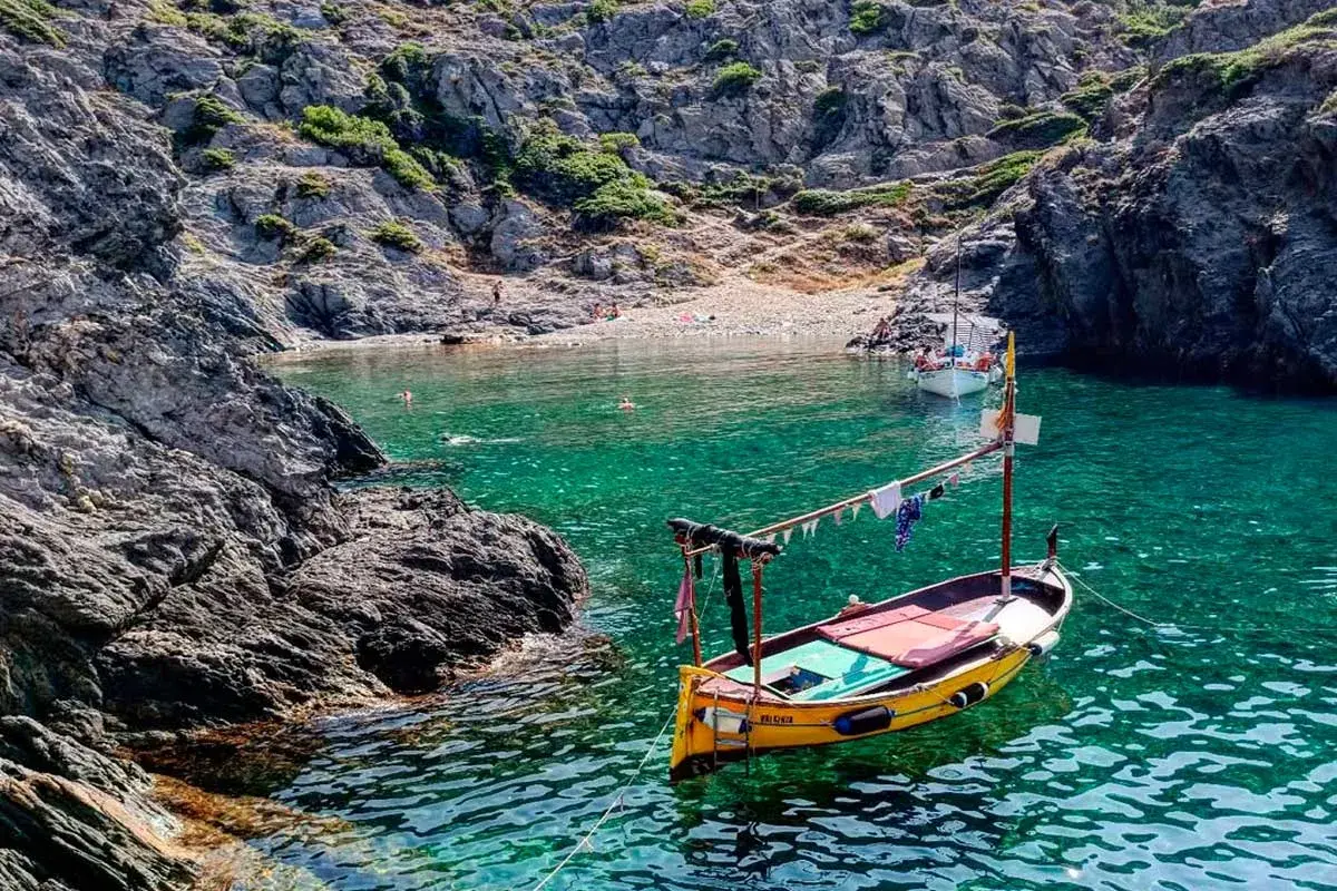 Barca amarilla en cala de aguas turquesas en el **Cabo de Creus**. Rocas escarpadas y gente en la playa al fondo.