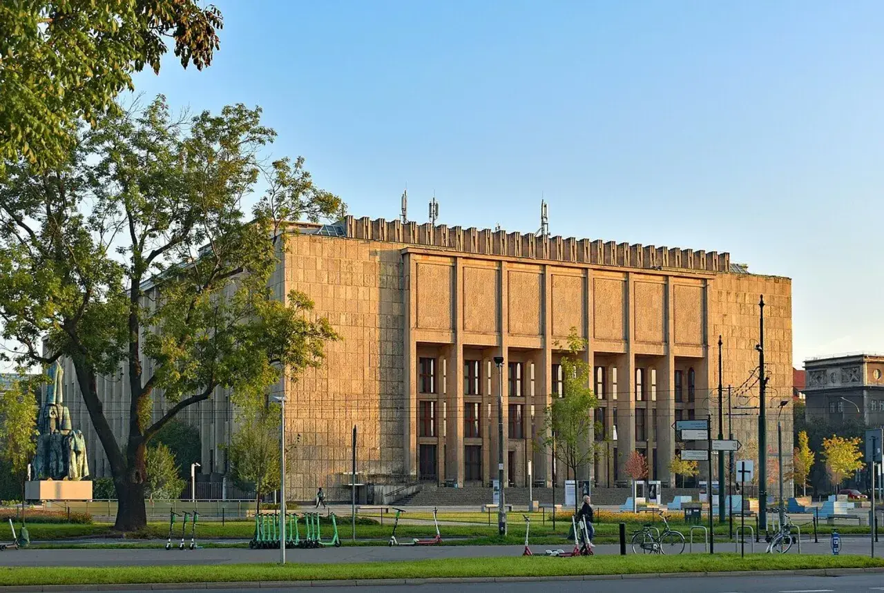 Muzeum Narodowe w Krakowie przy Alei 3 Maja. Monumentalna budowla z kamienia, otoczona zielenią i stojakami z hulajnogami.