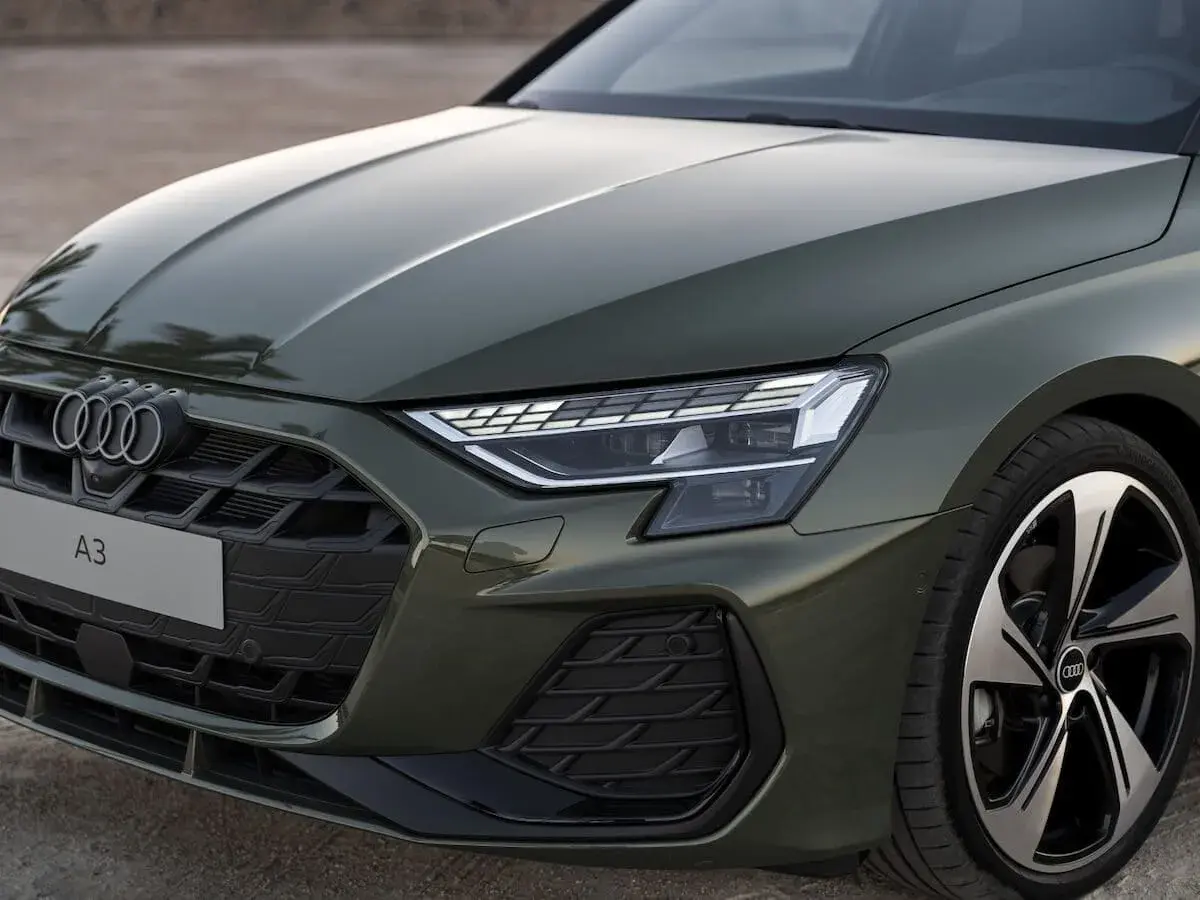 Nowe Audi A3 w zielonym kolorze prezentuje się elegancko. Widoczny fragment maski, grill z logo i reflektor podkreślają jego nowoczesny design.